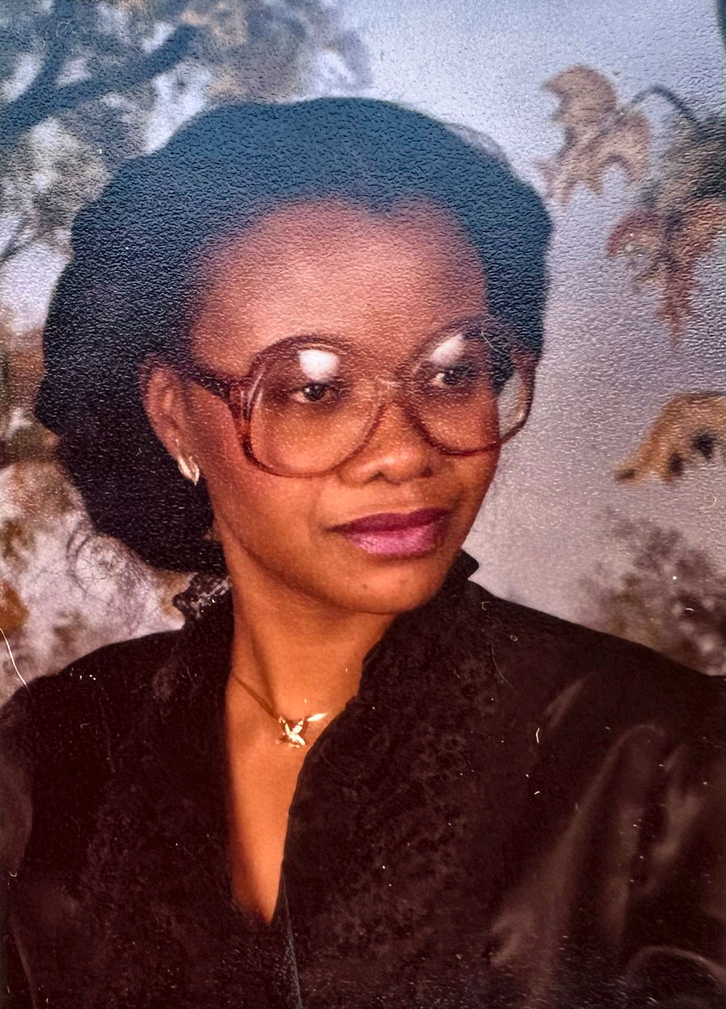 Archival portrait of a woman in round eyeglasses and a dark blouse with a delicate necklace, before a painted autumn backdrop.