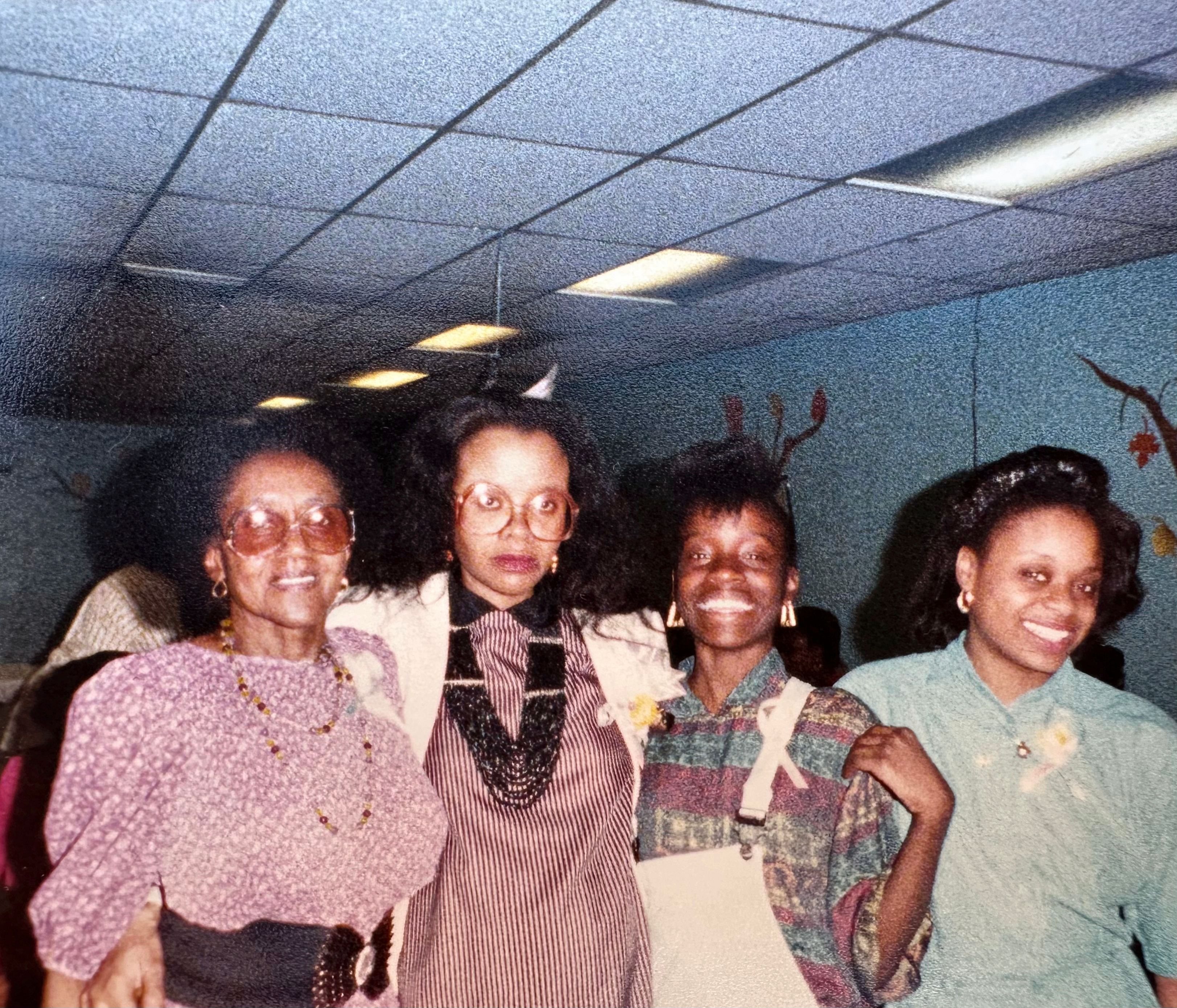 Four women standing together and smiling at a celebration beneath a tiled ceiling.