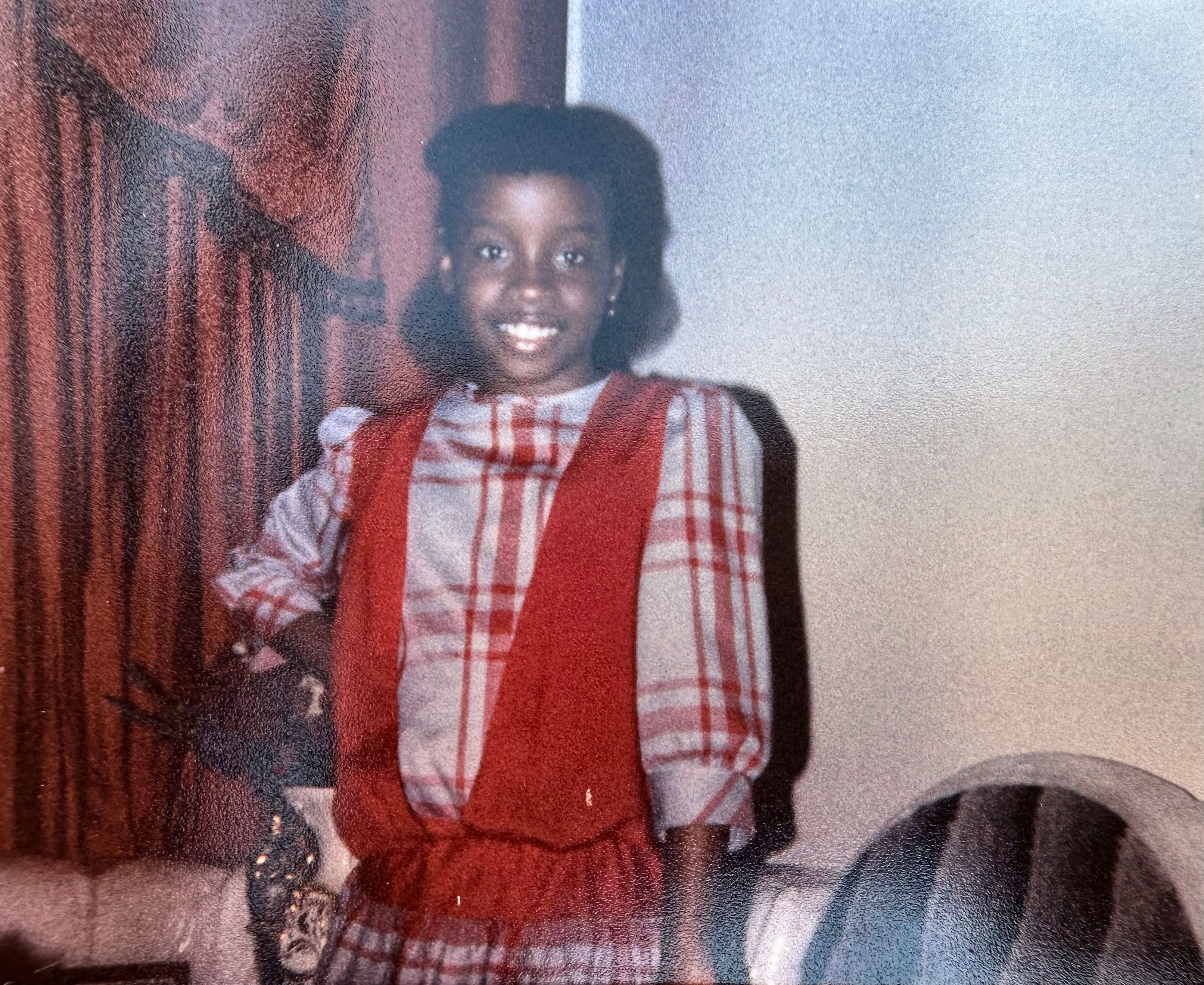 A smiling young girl in a plaid blouse and red vest stands in a living room.