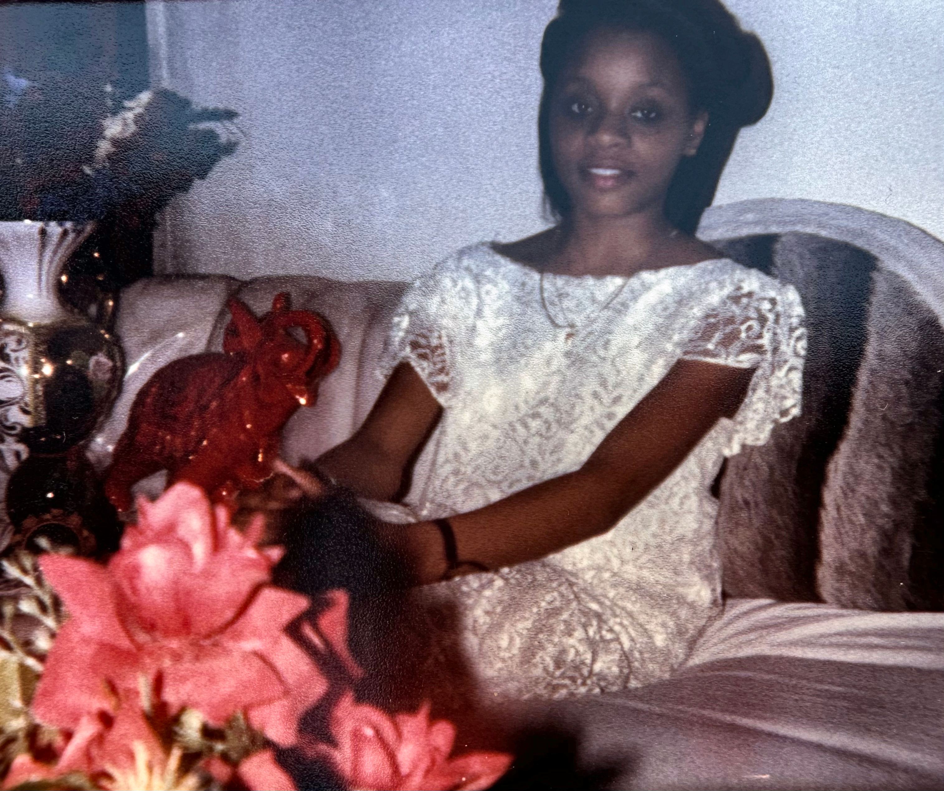 A young girl in a white lace dress seated on a sofa beside a vase of pink flowers.