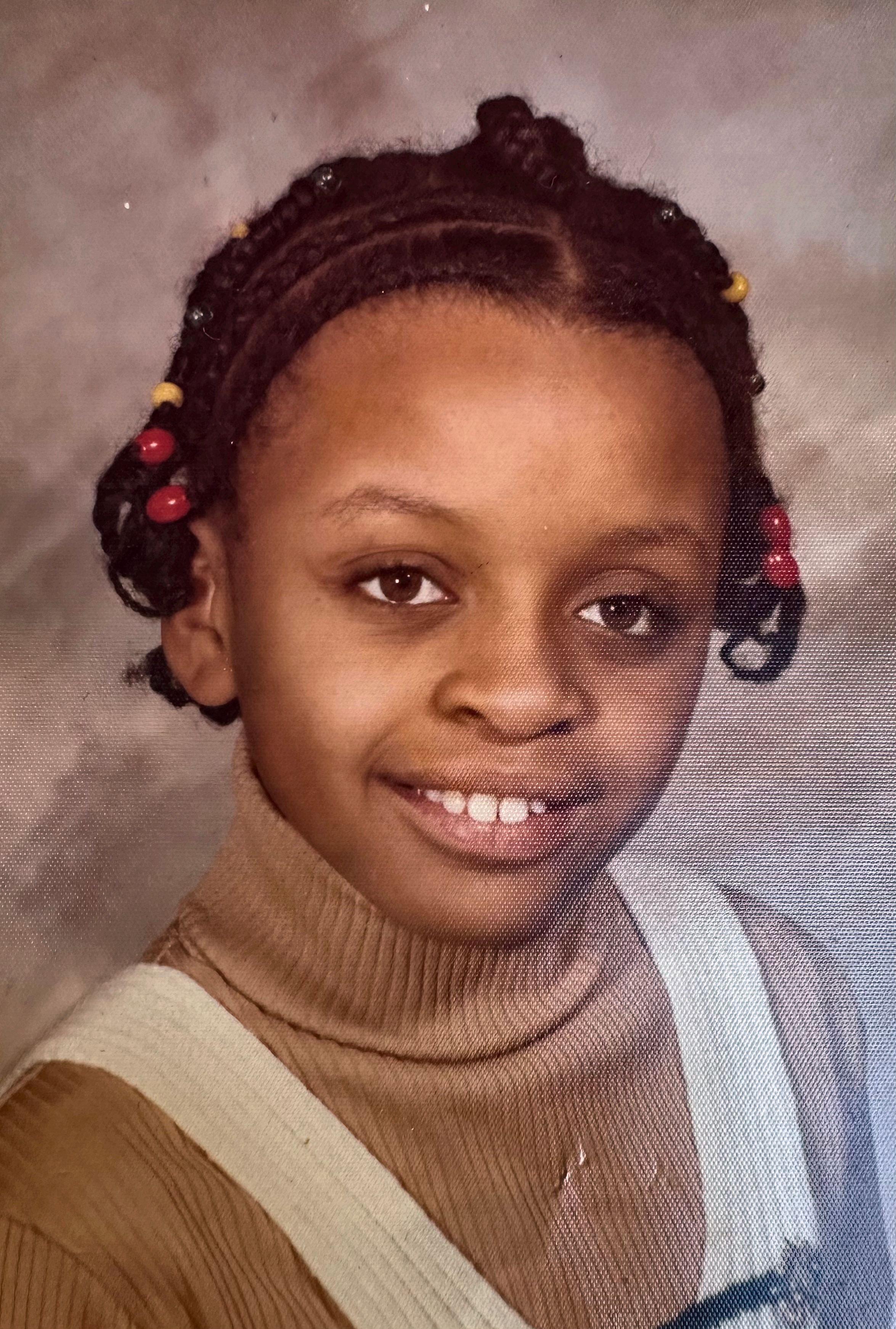 School portrait of a smiling young girl with braided hair and red beads, wearing a tan turtleneck.